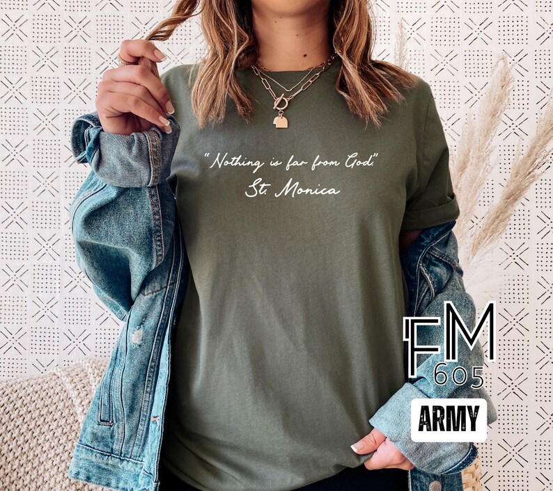 Puede incluir: Una camiseta verde con la cita "Nada est&aacute; lejos de Dios" y el nombre "Santa M&oacute;nica" escrito en blanco. La camiseta la lleva puesta una persona. La imagen tambi&eacute;n incluye una chaqueta vaquera y el texto "FM 605 ARMY".