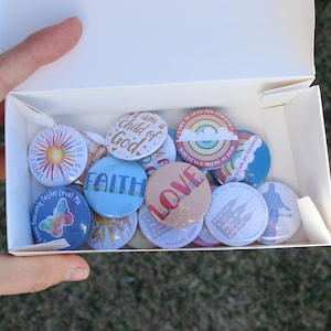 May include: A white box filled with colorful round buttons. The buttons feature various designs, including a sunburst, the word "FAITH", a butterfly, and the word "LOVE". The box is held by a hand, with a grassy background.