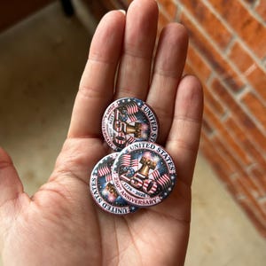 Puede incluir: Tres botones redondos con un diseño patriótico se sostienen en una mano. Los botones presentan el texto "UNITED STATES" y "250th Anniversary 1776-2026" con una bandera estadounidense y una representación de una maza.