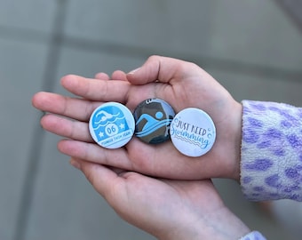 Op de afbeelding: Drie ronde buttons met zwemthema. Eén is lichtblauw met een zwemmer-icoon en de tekst 06 PIONEER SWIM TEAM. Een andere is donkerblauw met een zwemmer-icoon. De derde is wit met de tekst JUST KEEP SWIMMING.
