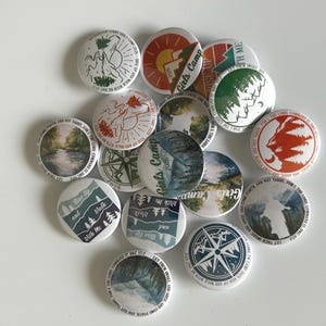 Puede incluir: Una colección de botones redondos y blancos con varios diseños. Algunos presentan escenas de montañas y bosques, brújulas y texto como "Girls Camp 2026" y "Rise Up and Walk With Me". La paleta de colores incluye verdes, azules y naranjas.