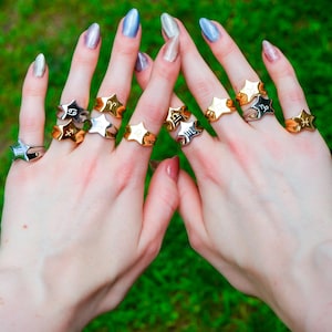 May include: A collection of star-shaped rings, each featuring a different zodiac symbol. The rings are in gold and silver tones, displayed on fingers against a green background. The rings are a unique fashion accessory.
