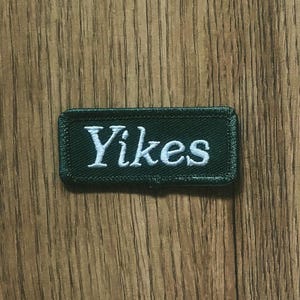 May include: A rectangular, dark green embroidered patch with the word "Yikes" in white, stylized lettering. The patch has a black border and is set against a wooden background.