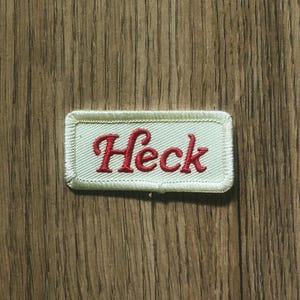 May include: A rectangular patch with the word "Heck" embroidered in red script on a white background. The patch has a light green border and is set against a wood grain surface.
