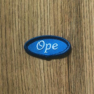 May include: An oval-shaped blue patch with the word "Ope" embroidered in white. The patch has a dark blue border and is set against a wooden background.