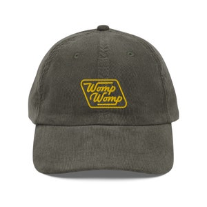 Può includere: Un cappello da baseball in velluto a coste verde scuro con un logo ricamato giallo che dice "Womp Womp" in un font retrò.