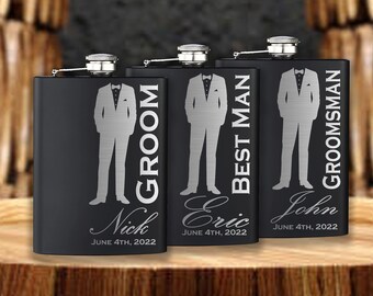Può includere: Tre fiaschette in metallo nero con testo inciso. Le fiaschette sono etichettate "Groom", "Best Man" e "Groomsman". Ogni fiaschetta ha un nome e una data diversi incisi.