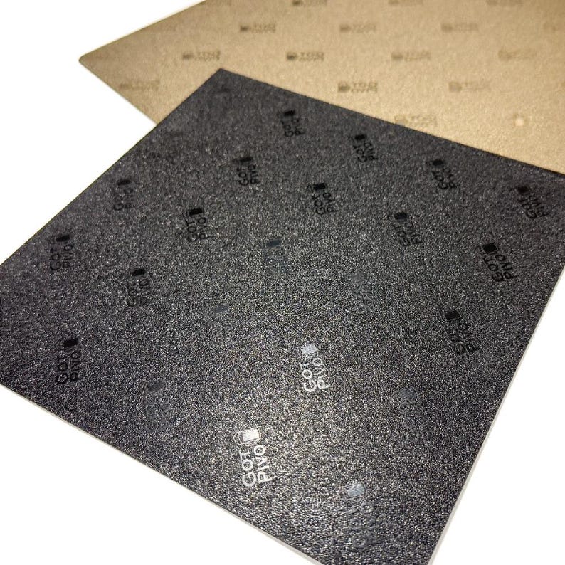 Pu&ograve; includere: Due fogli quadrati di materiale testurizzato, uno nero e uno beige, entrambi con un motivo ripetuto del testo "GOT Pivo" stampato in caratteri piccoli sulla superficie. I fogli sono sovrapposti, con quello nero posizionato sopra.