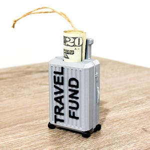 Puede incluir: Un peque&ntilde;o adorno de maleta gris con un billete de veinte d&oacute;lares enrollado en su interior. La maleta tiene las palabras "TRAVEL FUND" impresas en el lateral.