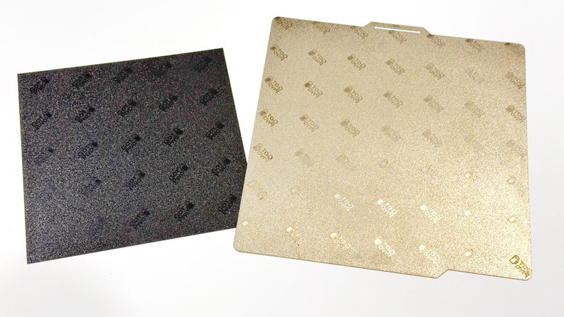 Pu&ograve; includere: Due piastre di costruzione quadrate per stampanti 3D. Una piastra &egrave; grigio scuro con una superficie ruvida e un motivo a logo ripetuto. L'altra piastra &egrave; color oro con finitura metallica, motivo a logo ripetuto e una piccola tacca.