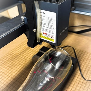 Pu&ograve; includere: Primo piano di un incisore laser con un raggio laser rosso focalizzato su un bicchiere trasparente. L'incisore &egrave; nero e ha un'etichetta bianca con il testo "XTOOL" e altre informazioni sulla societ&agrave; e sul prodotto. Il bicchiere &egrave; su una superficie marrone.
