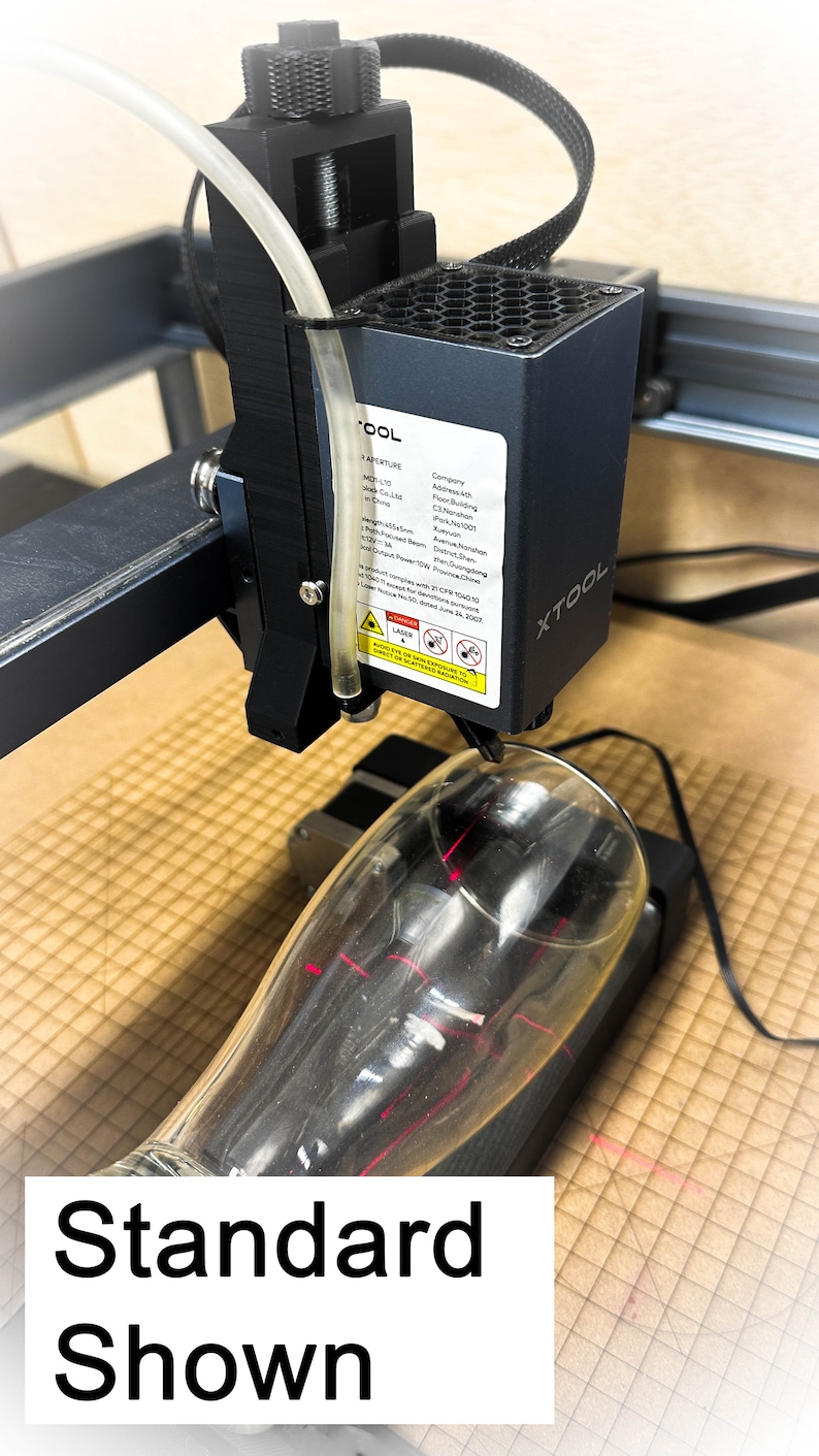 Pu&ograve; includere: Primo piano di un incisore laser con un raggio laser rosso focalizzato su un bicchiere trasparente. L'incisore &egrave; nero e ha un'etichetta bianca con il testo "XTOOL" e altre informazioni sulla societ&agrave; e sul prodotto. Il bicchiere &egrave; su una superficie marrone.