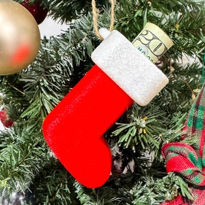 Puede incluir: Una media roja de fieltro con un pu&ntilde;o blanco cuelga de un &aacute;rbol de Navidad. Un billete de veinte euros est&aacute; metido dentro de la media.