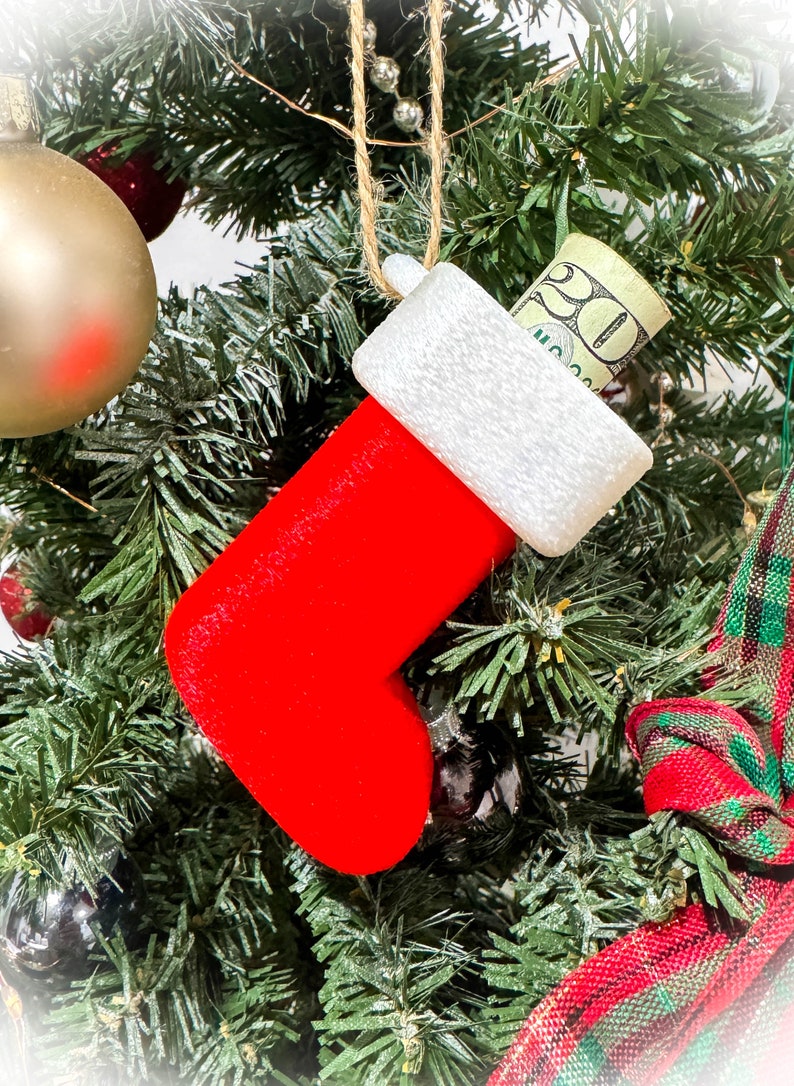 Puede incluir: Una media roja de fieltro con un pu&ntilde;o blanco cuelga de un &aacute;rbol de Navidad. Un billete de veinte euros est&aacute; metido dentro de la media.