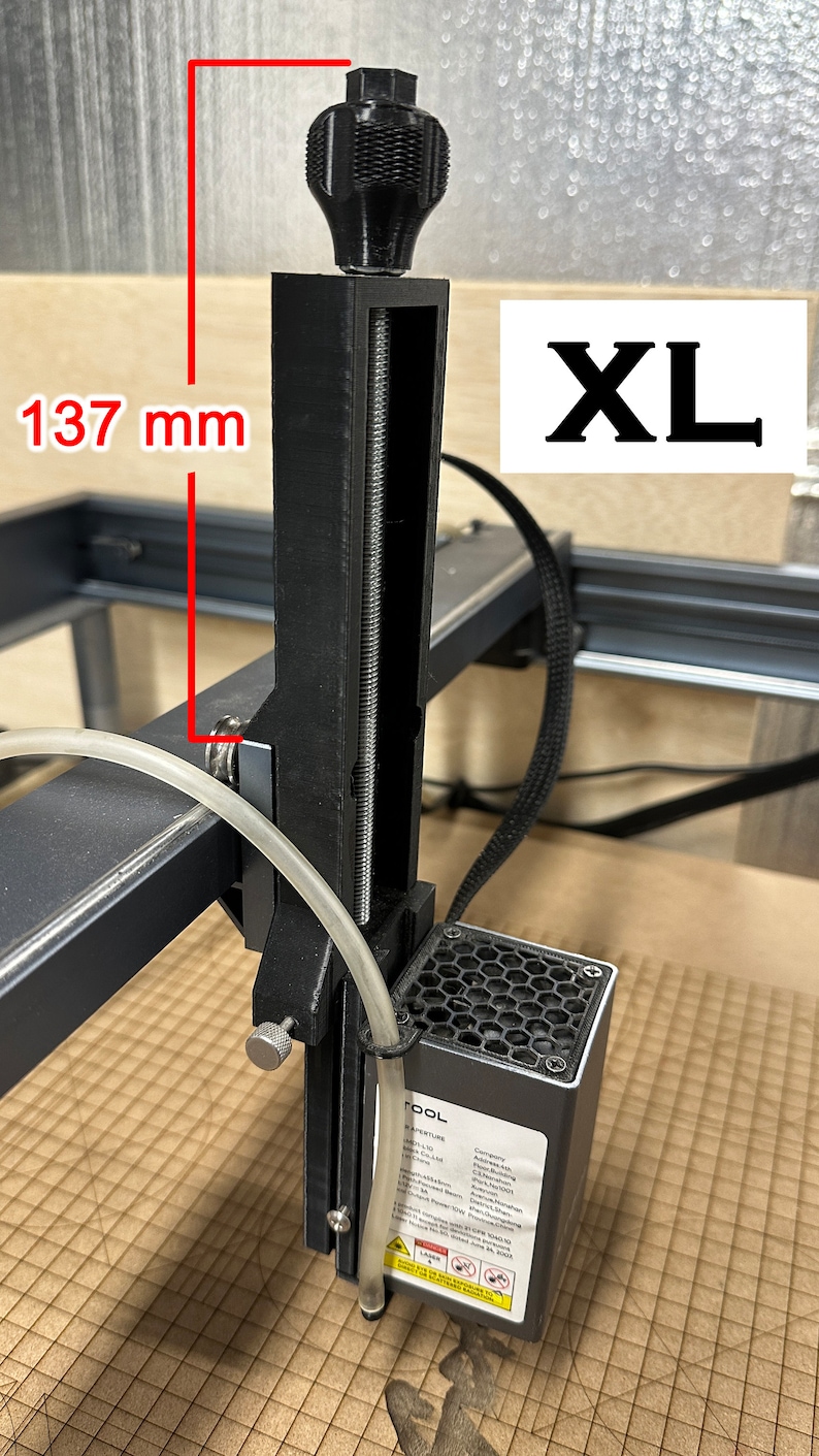 Pu&ograve; includere: Un estrusore per stampante 3D nero con una barra filettata argentata e una manopola nera. L'estrusore &egrave; montato su un telaio in metallo nero. L'estrusore &egrave; etichettato "TOOL" e ha un'etichetta di avvertenza sui laser. L'estrusore &egrave; alto 137 mm.