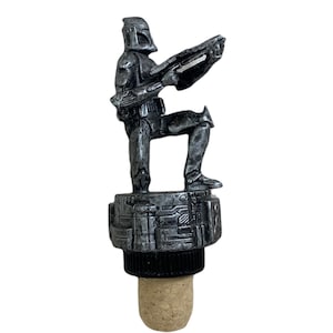 May include: A silver-coloured bottle stopper featuring a Star Wars clone trooper figurine holding a blaster rifle. The trooper is standing on a platform with a textured surface. The stopper has a cork base.