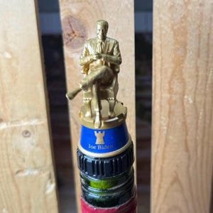 May include: A gold-colored bottle stopper featuring a seated figure. The stopper has a blue band with the text "Joe Biden" and a castle icon. The bottle stopper is on top of a wine bottle.