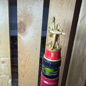 Op de afbeelding: Een decoratieve flesstopper met een goudkleurig beeld van een zittende man, bovenop een stapel flessendoppen. De stopper heeft een rode basis met gouden letters en een kasteelicoon.