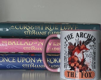 May include: White ceramic mug with a pink handle and rim, featuring a red fox illustration with floral accents and the text "THE ARCHER THE FOX." The mug is set in front of a stack of books with gold lettering.