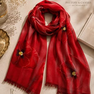 May include: A vibrant red scarf featuring a large-scale floral print with dark centers and yellow accents. The scarf has a semi-sheer, lightweight texture and finished edges with delicate fringe detailing at both ends. The text Pottery & Canvas is visible.