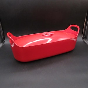 May include: A bright red rectangular casserole dish with a matching lid. The dish has two curved handles and a small knob on the lid. The overall design is simple and modern, suitable for baking and serving.