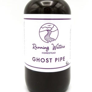 May include: A dark amber glass bottle with a black dropper top. The label reads "Running Waters Homestead" and "Ghost Pipe" in purple text. The bottle contains a dark liquid, likely a tincture or herbal remedy.