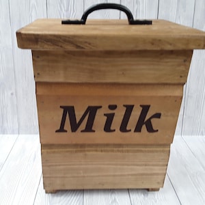 May include: A wooden milk box with a lid and a black handle. The word "Milk" is stencilled in dark brown on the front. The box is made of light brown wood.