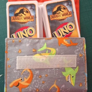 Może przedstawiać: Materiałowy uchwyt na karty z szarym zewnętrzem z wzorem pomarańczowych i zielonych ryb i gwiazd z kreskówek. Uchwyt zawiera dwa stosy kart UNO "Jurassic World". Uchwyt jest zabezpieczony białym zapięciem na rzep i ma pomarańczowe wnętrze.