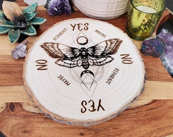 May include: A round wooden planchette with a moth design and the words "Yes", "No", "Maybe", and "Rephrase" etched into the wood. The planchette is surrounded by crystals, a candle, and a potted succulent.