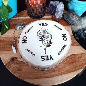 May include: A wooden planchette with a snake and moon design, surrounded by the words "Yes", "No", "Unclear", and "Rephrase". A lit candle in a mosaic glass holder sits nearby, along with crystals and a pendulum.