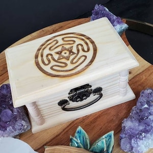 May include: A light-colored wooden box with a decorative etched design on the lid. The box has a metal clasp and handle. Amethyst crystals and a ceramic incense holder with a palo santo stick are also present.