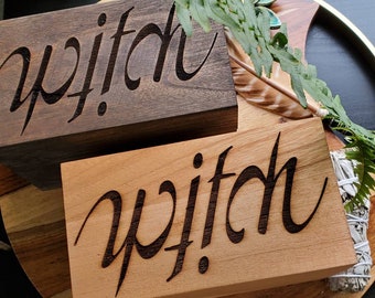 May include: Two wooden blocks with the word "witch" carved into them. One block is dark brown, the other is light brown. The blocks are on a wooden tray with greenery and a feather.