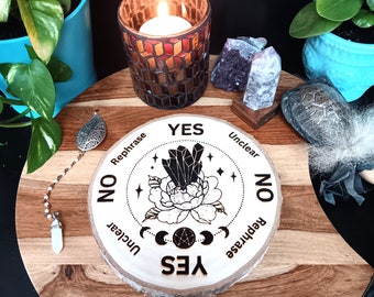 May include: A wooden divination board with the words "Yes", "No", "Unclear", and "Rephrase" encircling a floral and crystal design. A lit candle in a mosaic glass holder sits nearby, along with crystals and a pendulum.