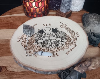 May include: A wooden planchette board with a carved design featuring two owls, Celtic knotwork, and the words "YES", "NO", "MAYBE", and "REPHRASE". Includes a silver leaf-shaped pendulum and a white crystal pendulum.