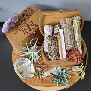 May include: A wooden box with the text "Sage that Shit!" and a bundle of sage, amethyst crystal, and other spiritual items. The box is open, revealing bundles of sage, palo santo, and a dried rose. The arrangement is on a wooden tray.