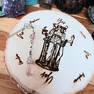 May include: A wooden divination board with a burned design of a three-faced figure and the word "Yes". A key-shaped pendulum with a pink crystal hangs above the board. The board is on a wooden surface.