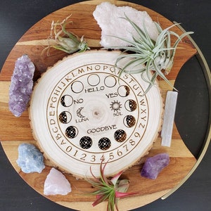 May include: A wooden planchette board with engraved letters, numbers, and moon phase symbols. The board is surrounded by crystals and air plants, creating a mystical aesthetic. The board includes the words "HELLO", "YES", "NO", "GOODBYE", "LUNA", and "SOL".