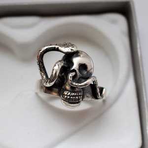 May include: A silver ring featuring a detailed skull with a snake wrapped around it. The snake's tail is curled around the skull's jaw.