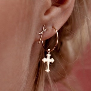 Blonde model's ear featuring thin solid gold hoop earrings with cross pendant