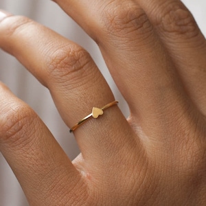May include: A delicate gold ring with a small heart-shaped design. The ring is worn on a finger.
