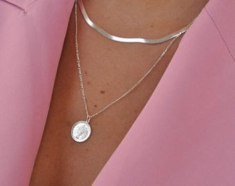 Op de afbeelding: Een gelaagde zilveren ketting set bestaande uit een platte, gladde slangenketting en een fijne schakelketting met een ronde muntvormige hanger met een reliëf van een paard, gedragen over een lichtroze blazer met reverskraag.