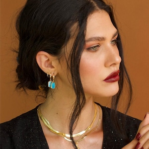 May include: A gold-toned layered herringbone chain necklace, a pair of gold hoop earrings with small rectangular turquoise-colored pendants, and a thin gold ring with a small white stone accent, worn with a black textured top.