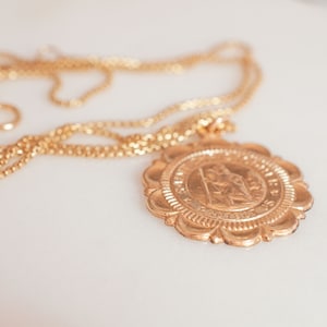 Peut inclure: Un collier en cha&icirc;ne dor&eacute;e avec un grand pendentif rond orn&eacute; d'un motif d&eacute;taill&eacute;. Le pendentif a une surface textur&eacute;e en relief avec une image centrale et une inscription.