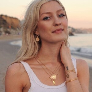 Peut inclure: Une femme aux longs cheveux blonds portant un d&eacute;bardeur blanc et des colliers en or avec diff&eacute;rents pendentifs, dont un papillon, une fleur et une pi&egrave;ce de monnaie. Elle porte &eacute;galement des boucles d'oreilles cr&eacute;oles en or et un bracelet en or avec un charme en forme de pi&egrave;ce de monnaie. La femme a un tatouage sur son poignet qui dit "808".