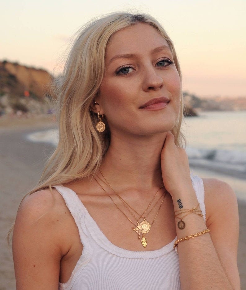 Peut inclure: Une femme aux longs cheveux blonds portant un d&eacute;bardeur blanc et des colliers en or avec diff&eacute;rents pendentifs, dont un papillon, une fleur et une pi&egrave;ce de monnaie. Elle porte &eacute;galement des boucles d'oreilles cr&eacute;oles en or et un bracelet en or avec un charme en forme de pi&egrave;ce de monnaie. La femme a un tatouage sur son poignet qui dit "808".