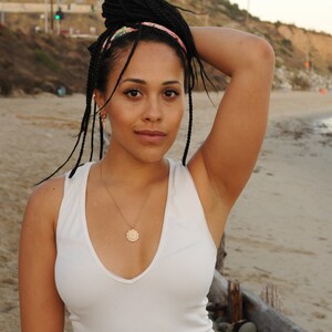 Peut inclure: Une femme avec de longues tresses noires portant une combinaison blanche sans manches et un collier en or avec un pendentif rond. Elle se tient sur une plage avec un rivage rocheux en arri&egrave;re-plan.