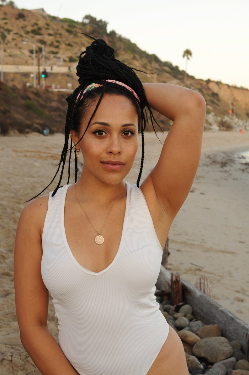 Peut inclure: Une femme avec de longues tresses noires portant une combinaison blanche sans manches et un collier en or avec un pendentif rond. Elle se tient sur une plage avec un rivage rocheux en arri&egrave;re-plan.