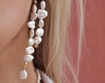 May include: A pair of long drop earrings featuring multiple irregular white baroque pearls strung together with small gold-toned metal beads. The earrings are attached to gold-toned posts and are worn alongside a matching white pearl necklace.
