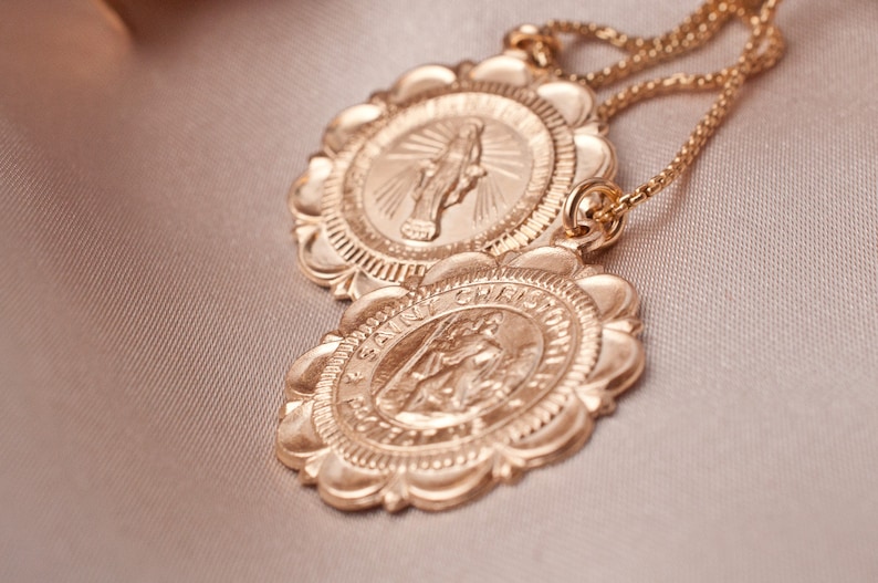Peut inclure: Deux m&eacute;daillons dor&eacute;s sur un collier &agrave; cha&icirc;ne. Les m&eacute;daillons sont ronds avec des bords festonn&eacute;s et pr&eacute;sentent des images religieuses. Le m&eacute;daillon sup&eacute;rieur repr&eacute;sente une femme avec les mains lev&eacute;es en pri&egrave;re. Le m&eacute;daillon inf&eacute;rieur repr&eacute;sente un homme avec une aur&eacute;ole et une croix. Le texte "SAINT CHRISTOPHER" est visible autour du bord du m&eacute;daillon inf&eacute;rieur.