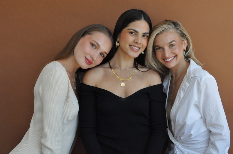 Peut inclure: Trois femmes portant des colliers et des boucles d'oreilles en or avec des pierres turquoise. La femme au milieu porte un haut noir &agrave; &eacute;paules d&eacute;nud&eacute;es, tandis que les femmes de chaque c&ocirc;t&eacute; portent des hauts blancs.